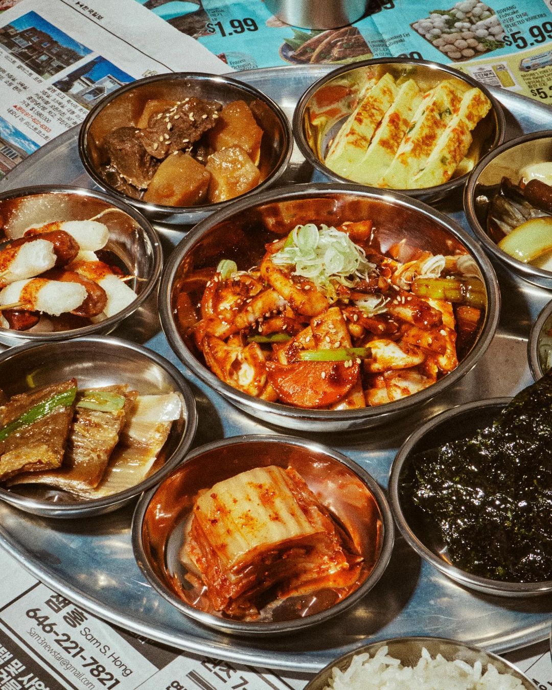 Overhead shot of a round metal tray with small bowls of Korean side dishes, including kimchi, seaweed, and marinated meats and vegetables.