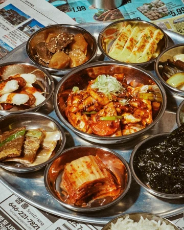 Overhead shot of a round metal tray with small bowls of Korean side dishes, including kimchi, seaweed, and marinated meats and vegetables.