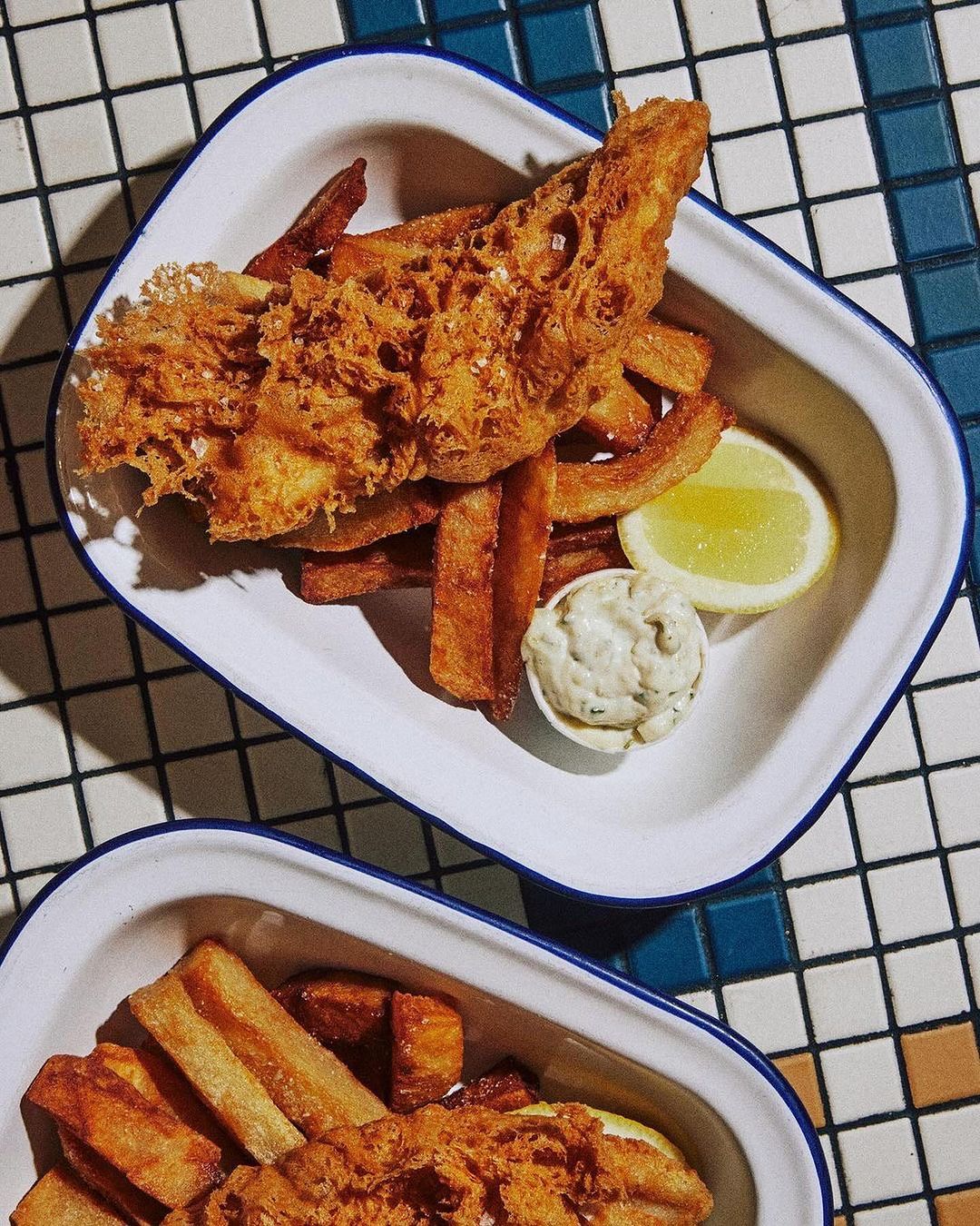 Overhead shot of fried fish, thick-cut fries, lemon wedge, and tartar sauce on a white plate with blue trim, set on a checkered surface.