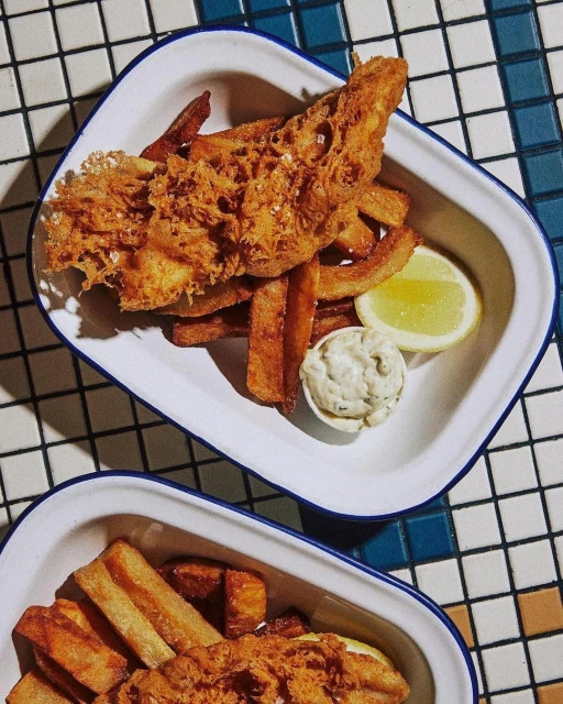 Overhead shot of fried fish, thick-cut fries, lemon wedge, and tartar sauce on a white plate with blue trim, set on a checkered surface.