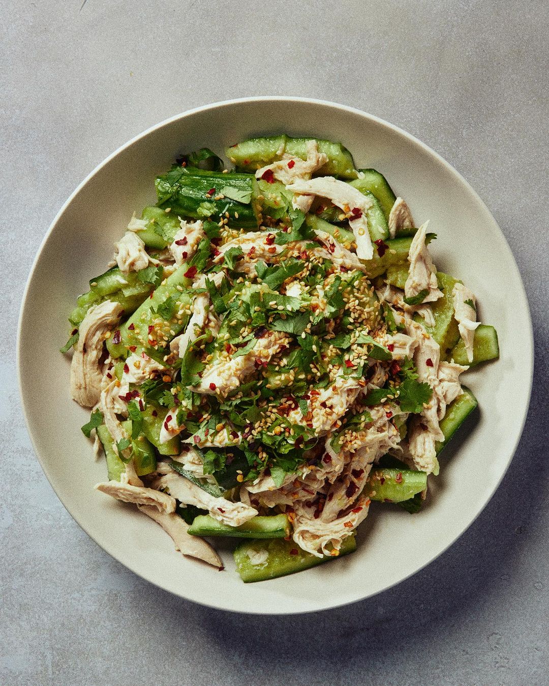 Overhead shot of a fresh cucumber and shredded chicken salad with cilantro and sesame seeds, presented in a white ceramic bowl.
