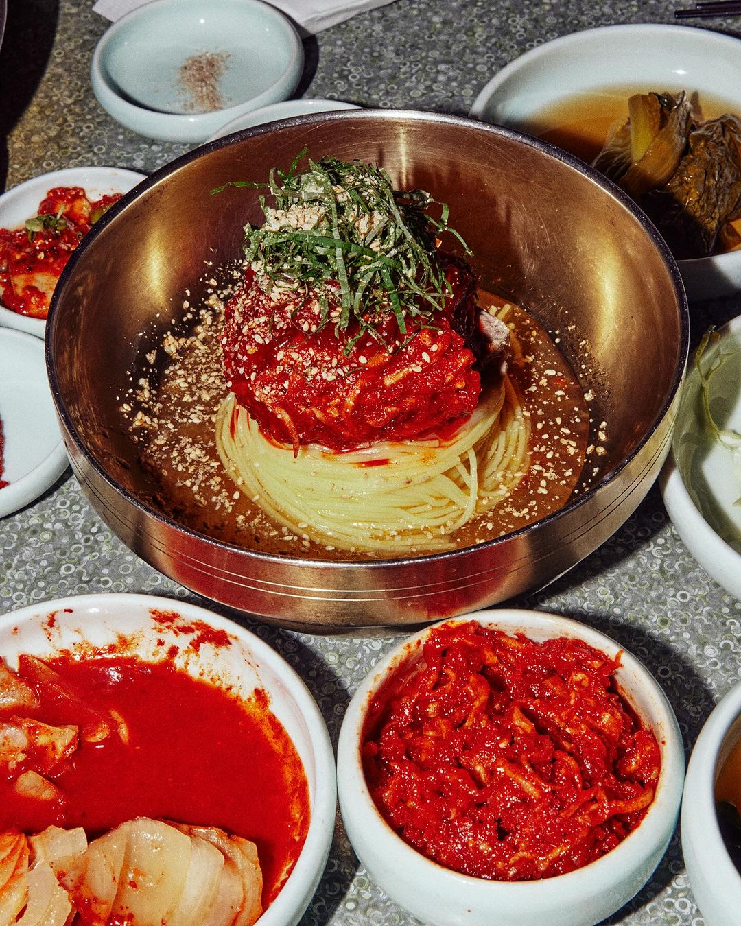 Korean cold noodles, known as naengmyeon, in a brass bowl with side dishes of kimchi, featuring a close-up, high-angle view.