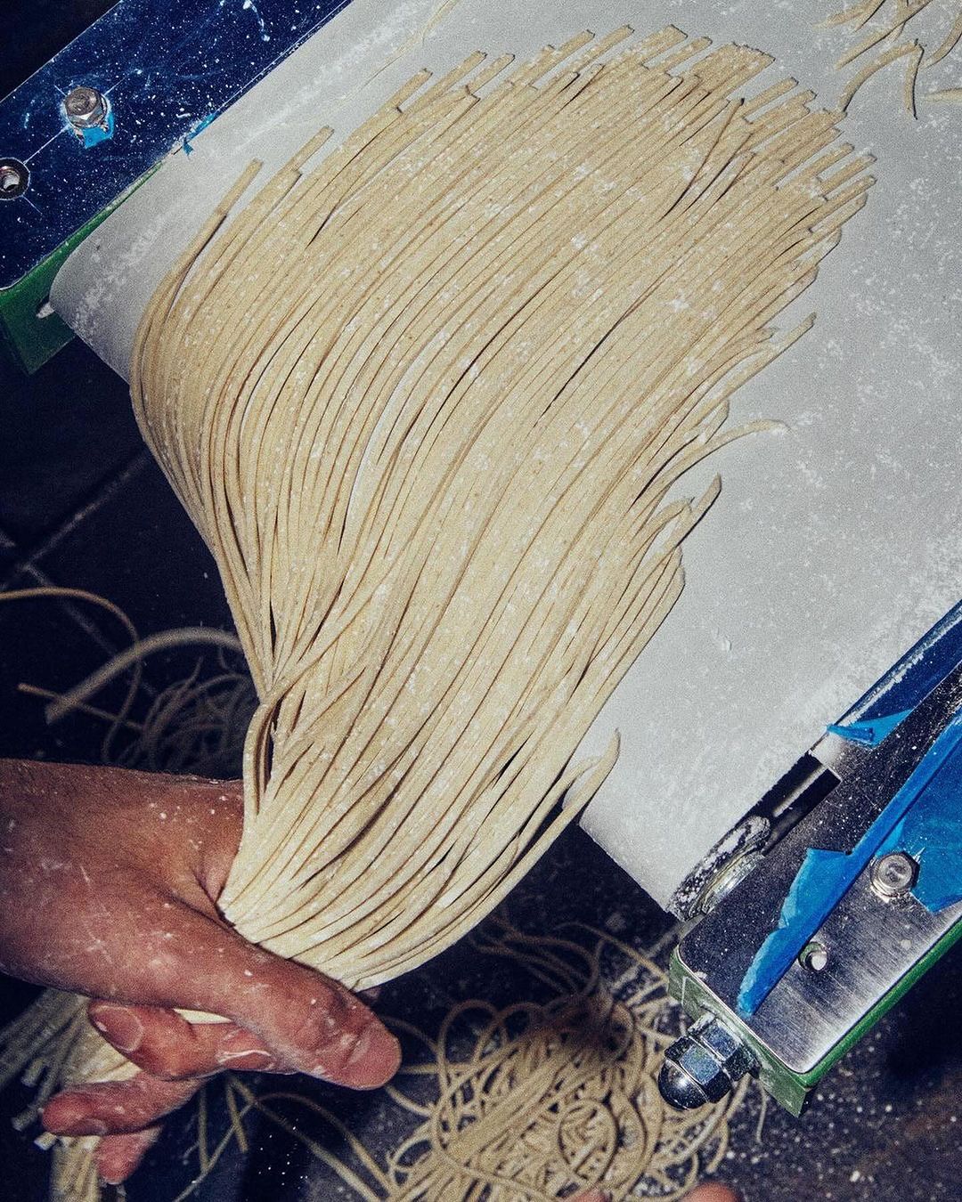 A close-up showcases long, light brown noodles being held by a hand, appearing to be processed by a food-making machine.