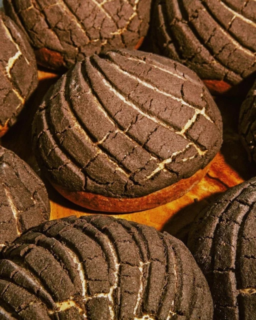 Close-up of a collection of freshly baked, dark brown concha sweet bread rolls with a ridged shell pattern on a wooden surface.