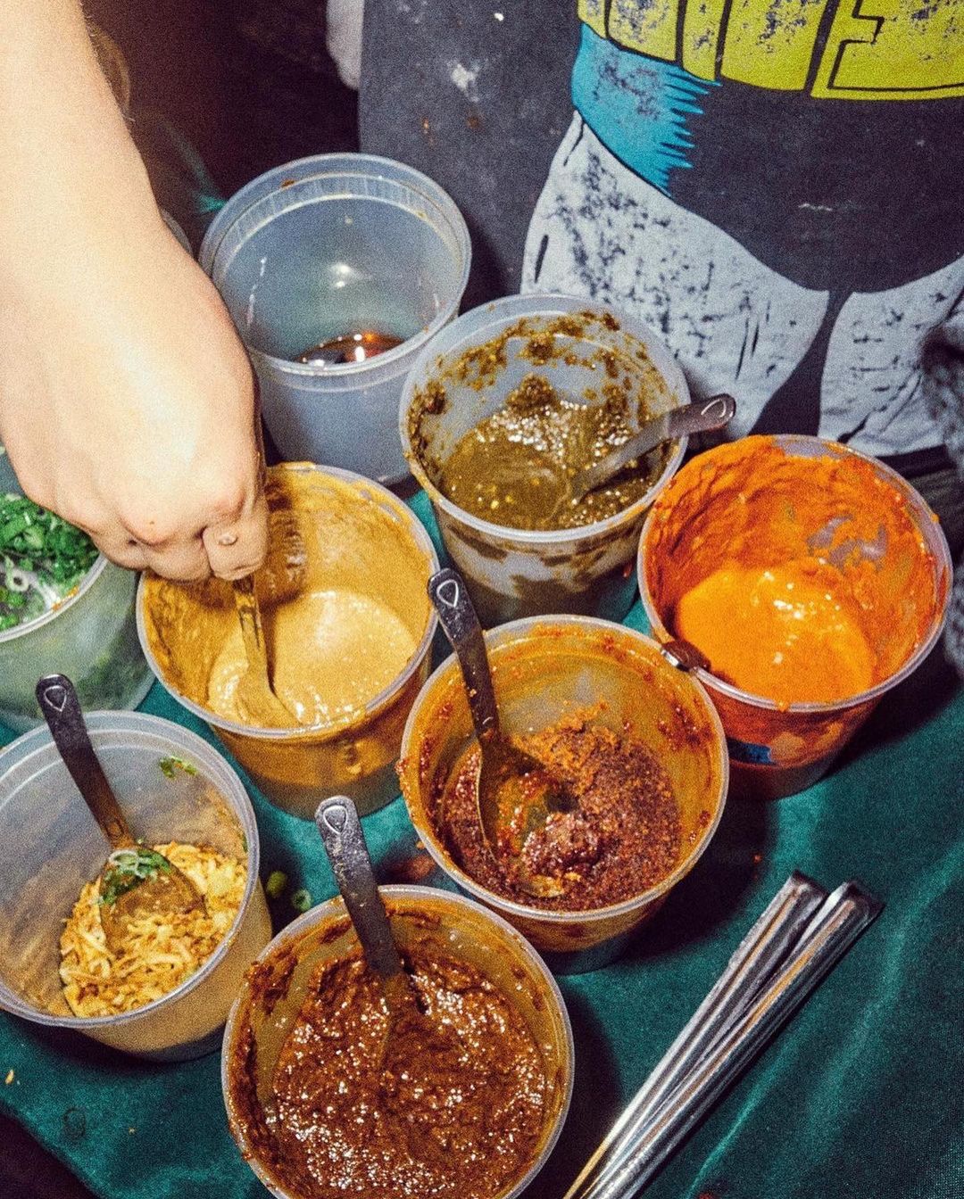 Overhead shot of various sauces in clear plastic containers, each with a spoon, alongside serving chopsticks.