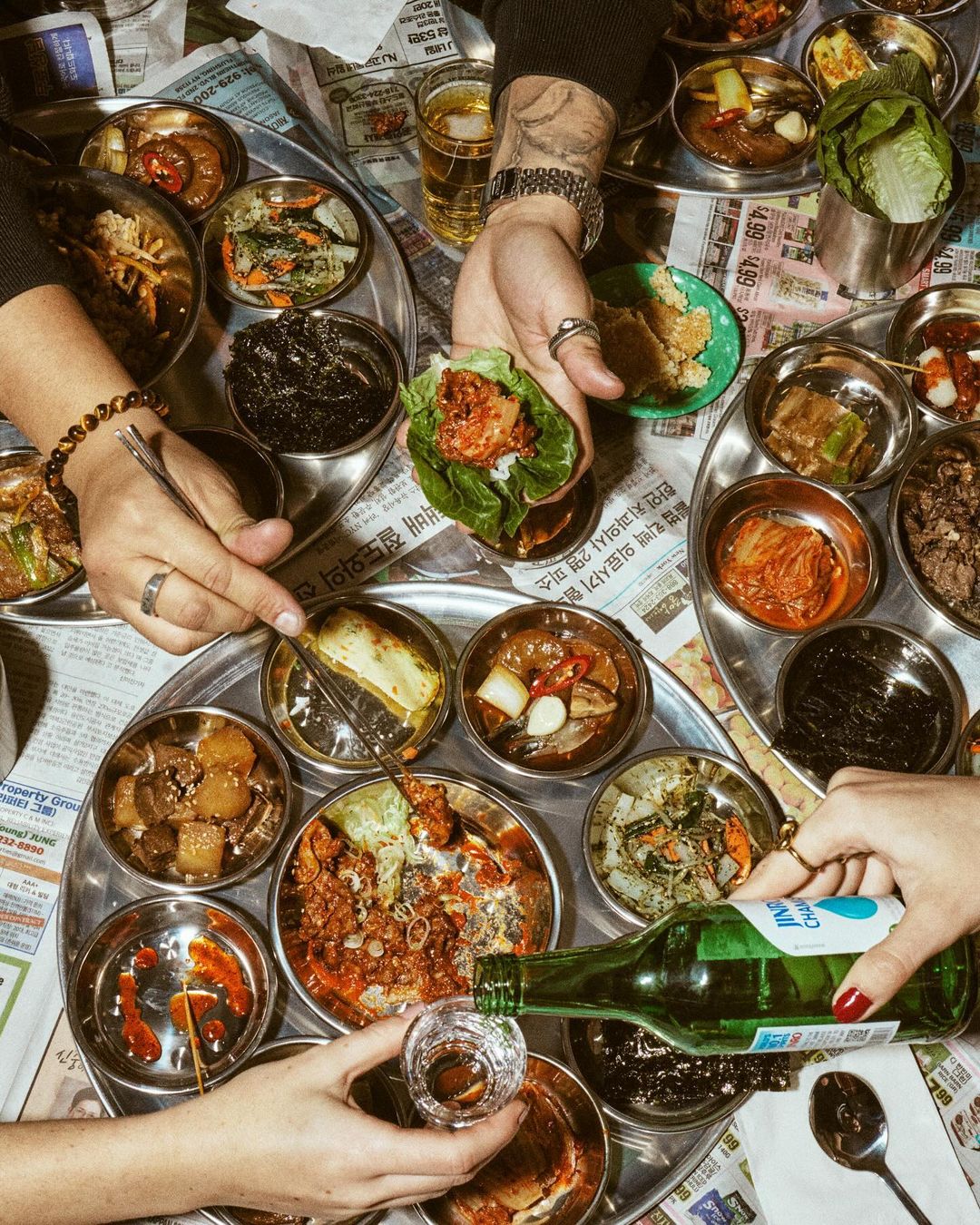 Overhead shot of hands arranging and pouring alcohol over an assortment of Korean dishes served on metal trays and newspaper.