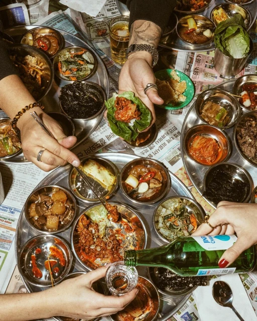 Overhead shot of hands arranging and pouring alcohol over an assortment of Korean dishes served on metal trays and newspaper.