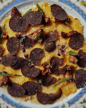 Overhead close-up of a white plate holding pasta dish with yellow sauce, black truffles, green herbs, and small mushrooms.