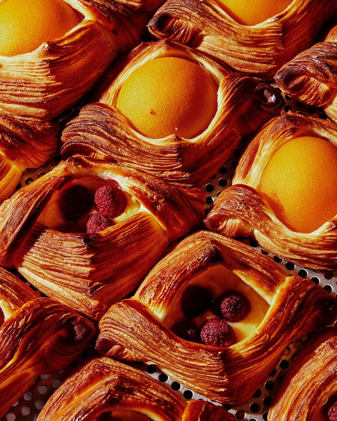 Close-up of assorted danishes on a metal rack: peach and raspberry fillings, golden-brown flaky crusts, glossy glaze in warm light.