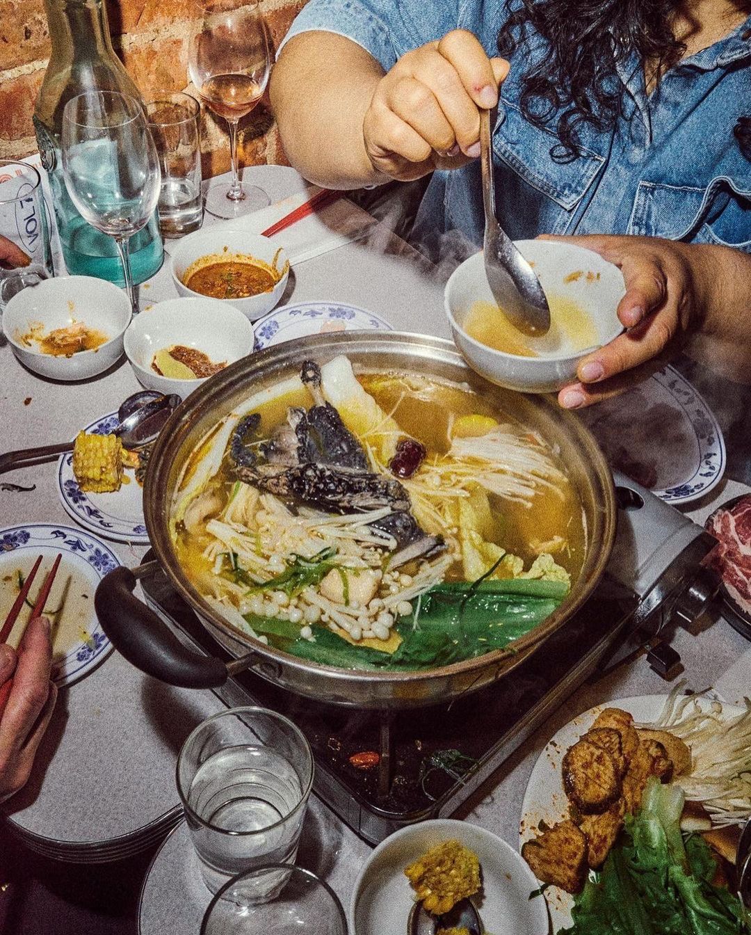 Overhead shot of a person ladling soup from a large pot surrounded by plates of food, drinks, and dishes in a warm-toned indoor setting.