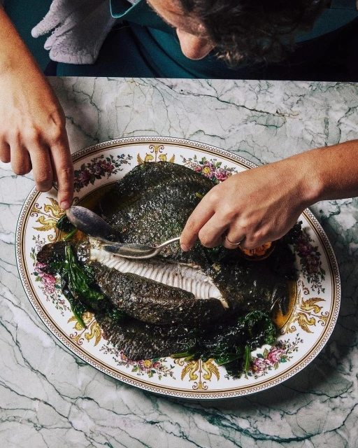 Overhead shot of a cooked fish being served on a floral-patterned plate, prepared by a person at a marble-top table.