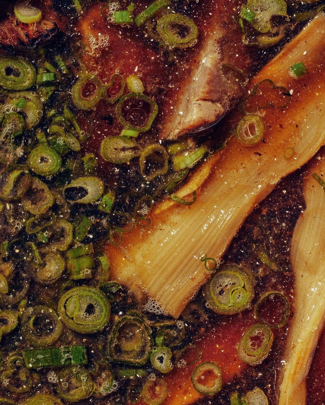 Overhead close-up of simmering broth with cuts of light brown meat, green onion scallions, and dark red-brown broth bubbling in a pot.