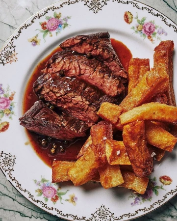 Overhead shot of sliced steak with brown sauce and a side of thick-cut fries on a decorative floral plate, resting on a marble surface.
