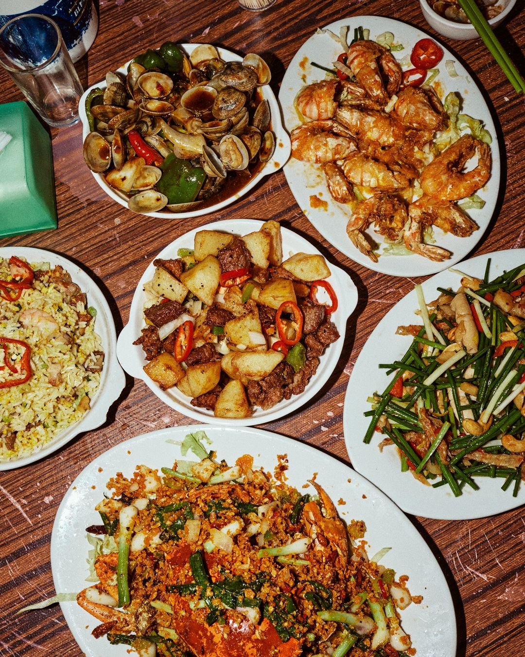 Overhead shot of a wooden table with a variety of cooked seafood dishes, including clams, prawns, and stir-fried vegetables.