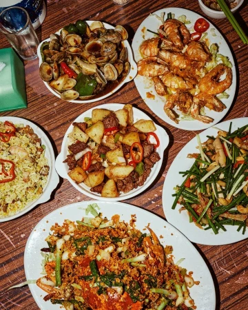 Overhead shot of a wooden table with a variety of cooked seafood dishes, including clams, prawns, and stir-fried vegetables.