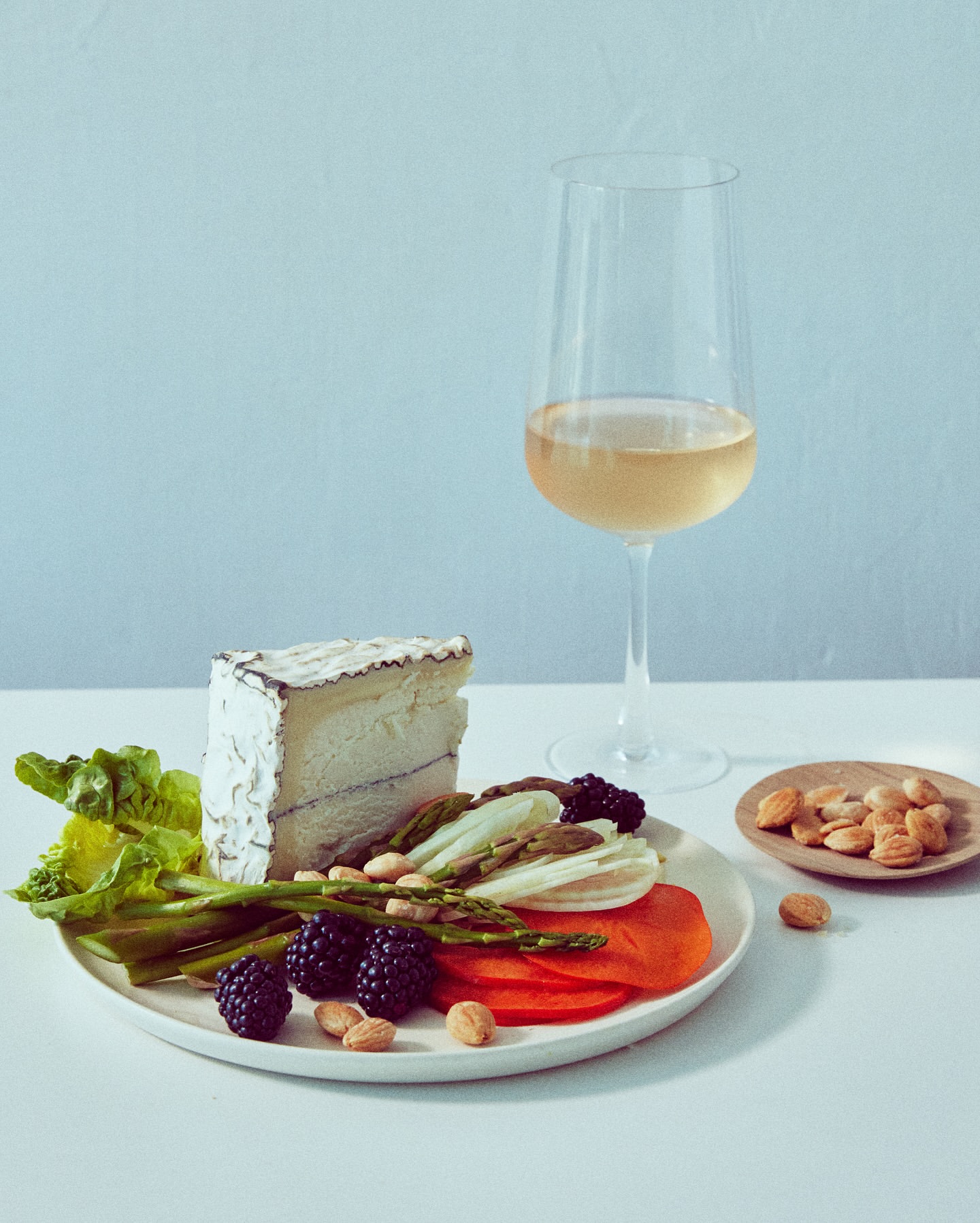 Cheese, sliced persimmons, blackberries, almonds and asparagus on a plate with white wine, creating a sophisticated food still life.