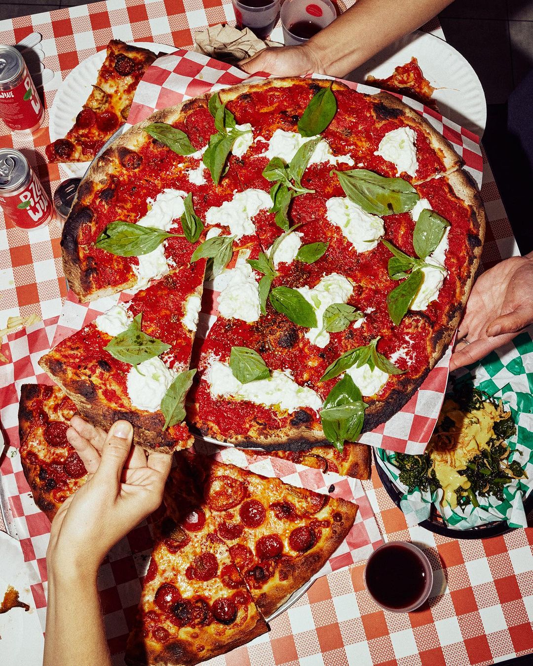 Overhead shot of a pizza topped with tomato sauce, mozzarella, and basil on a red and white checkered tablecloth, with hands and drinks.