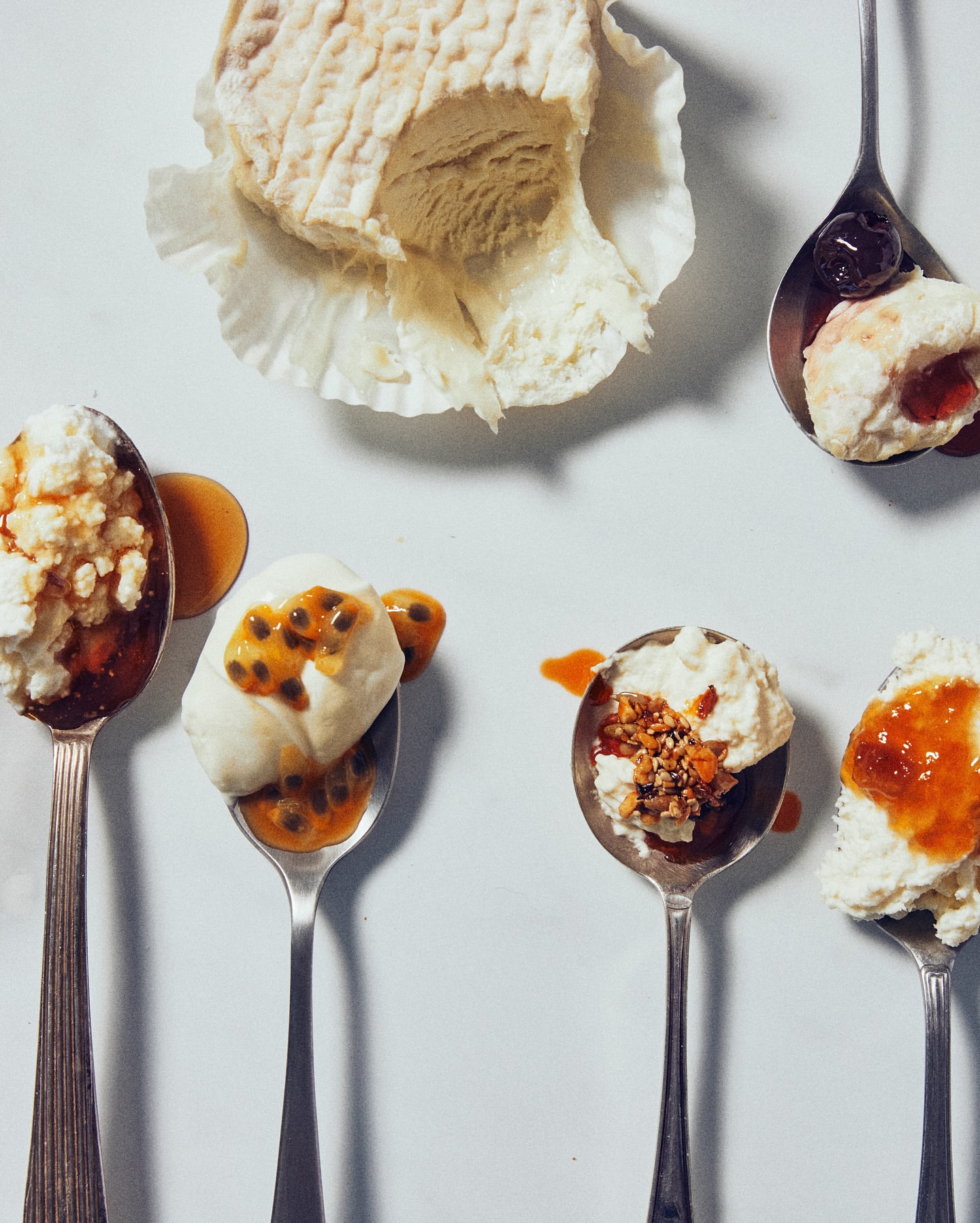 Overhead studio shot of a gourmet cheese block and spoons of desserts with fruit toppings, set against a bright white backdrop.