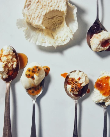 Overhead studio shot of a gourmet cheese block and spoons of desserts with fruit toppings, set against a bright white backdrop.