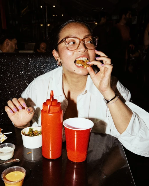 Woman in glasses eating food indoors at a table with ketchup bottle and red cup, bright light and dark background.