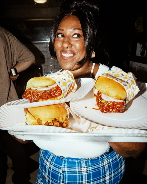 Woman smiling with three sandwiches on plates. She wears a plaid skirt, and the sandwiches are wrapped in patterned paper.