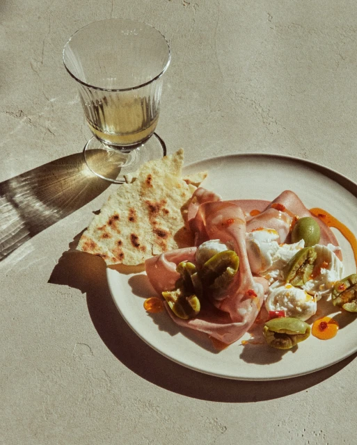 Overhead shot of mortadella, mozzarella, olives, and flatbread on a plate, accompanied by a wine glass, set against a neutral backdrop.