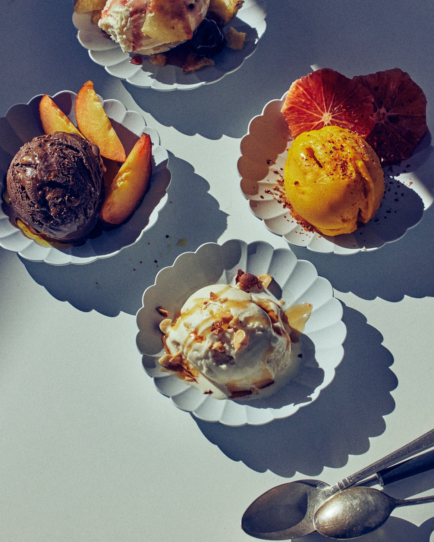 Overhead shot of four dessert plates, each with a scoop of ice cream and fruit slices. Spoons are in the lower right corner.