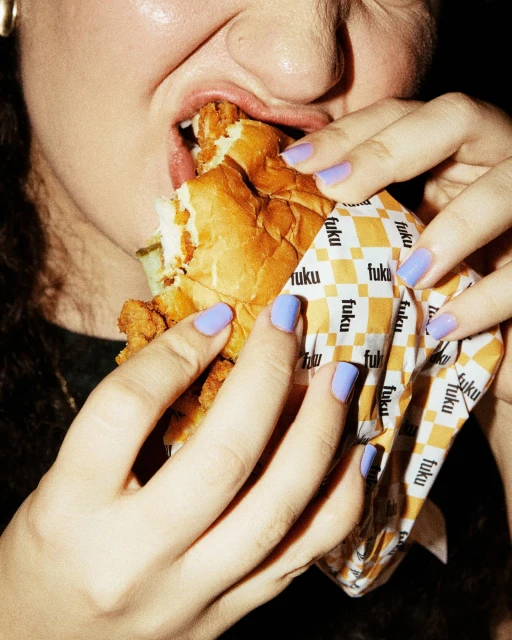 Close-up of an adult eating a chicken sandwich held in a wrapper, with light-purple painted fingernails. The background is dark.
