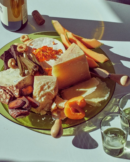 A vibrant green platter displays an assortment of cheeses, fruits, and chocolates next to a bottle of white wine, set in bright, natural light.