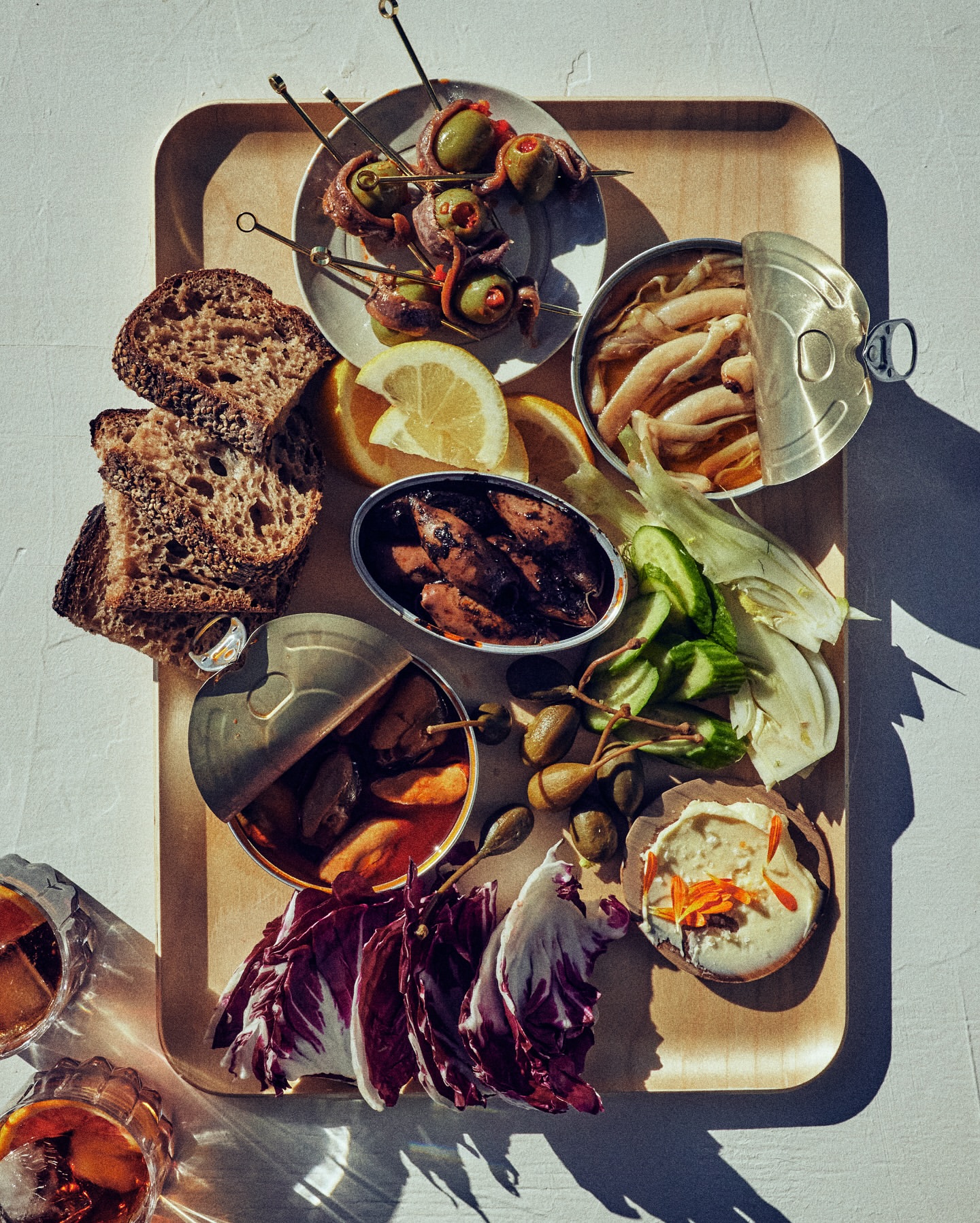 Overhead shot of a wooden tray filled with canned seafood, bread, olives, capers, lemon wedges, and cocktails, on a white surface.