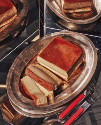 Sliced caramel flan served on an antique silver platter with serving utensils next to mirrored surfaces.