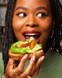 Close-up portrait of a Black woman eating an open-faced sandwich topped with greens and sliced fruit, photographed by Alex Lau.