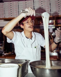 Chef in white apron and cap stretching warm mozzarella cheese with metal tongs in a bustling kitchen by Alex Lau