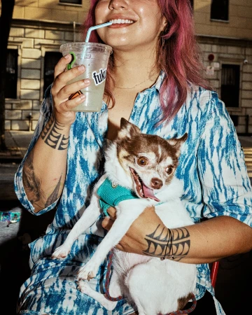 Woman with pink-dyed hair wearing a blue and white patterned shirt holding a small white and brown dog and a clear drink cup