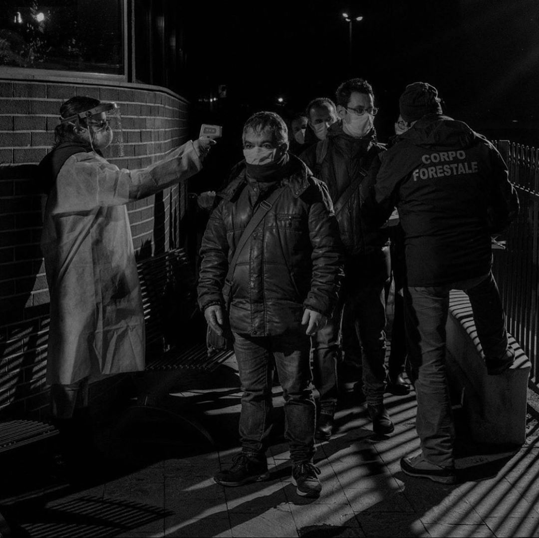 A group of people wearing face masks wait in line at night while a healthcare worker takes temperatures with a thermometer.