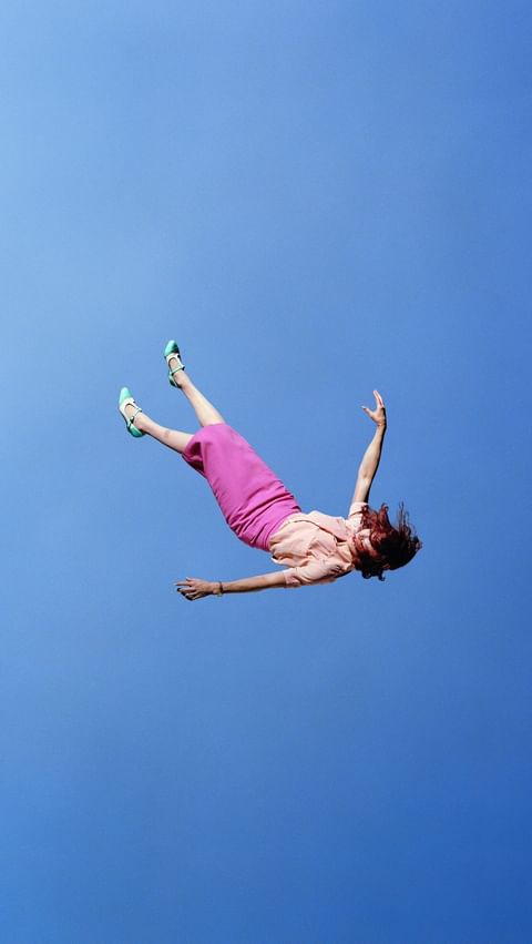 Woman in mid-air falling or floating with a pink skirt, light peach blouse, and blue shoes against a clear gradient blue sky.