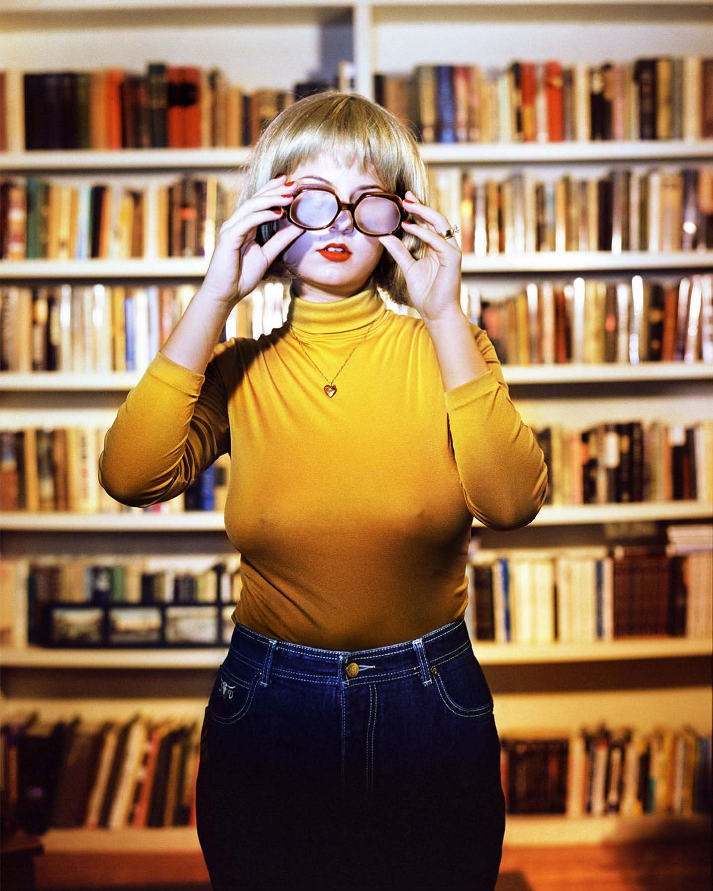 Woman wearing yellow turtleneck and blue jeans adjusting glasses in front of a filled bookshelf