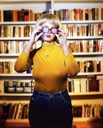 Woman wearing yellow turtleneck and blue jeans adjusting glasses in front of a filled bookshelf