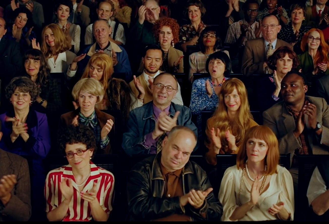 Diverse audience clapping in tiered seating: men and women of various ages and ethnicities, in bright lighting, creating a lively atmosphere.