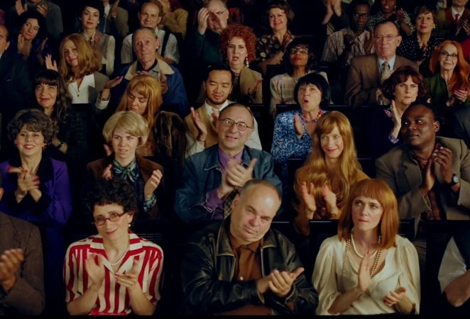 Diverse audience clapping in tiered seating: men and women of various ages and ethnicities, in bright lighting, creating a lively atmosphere.