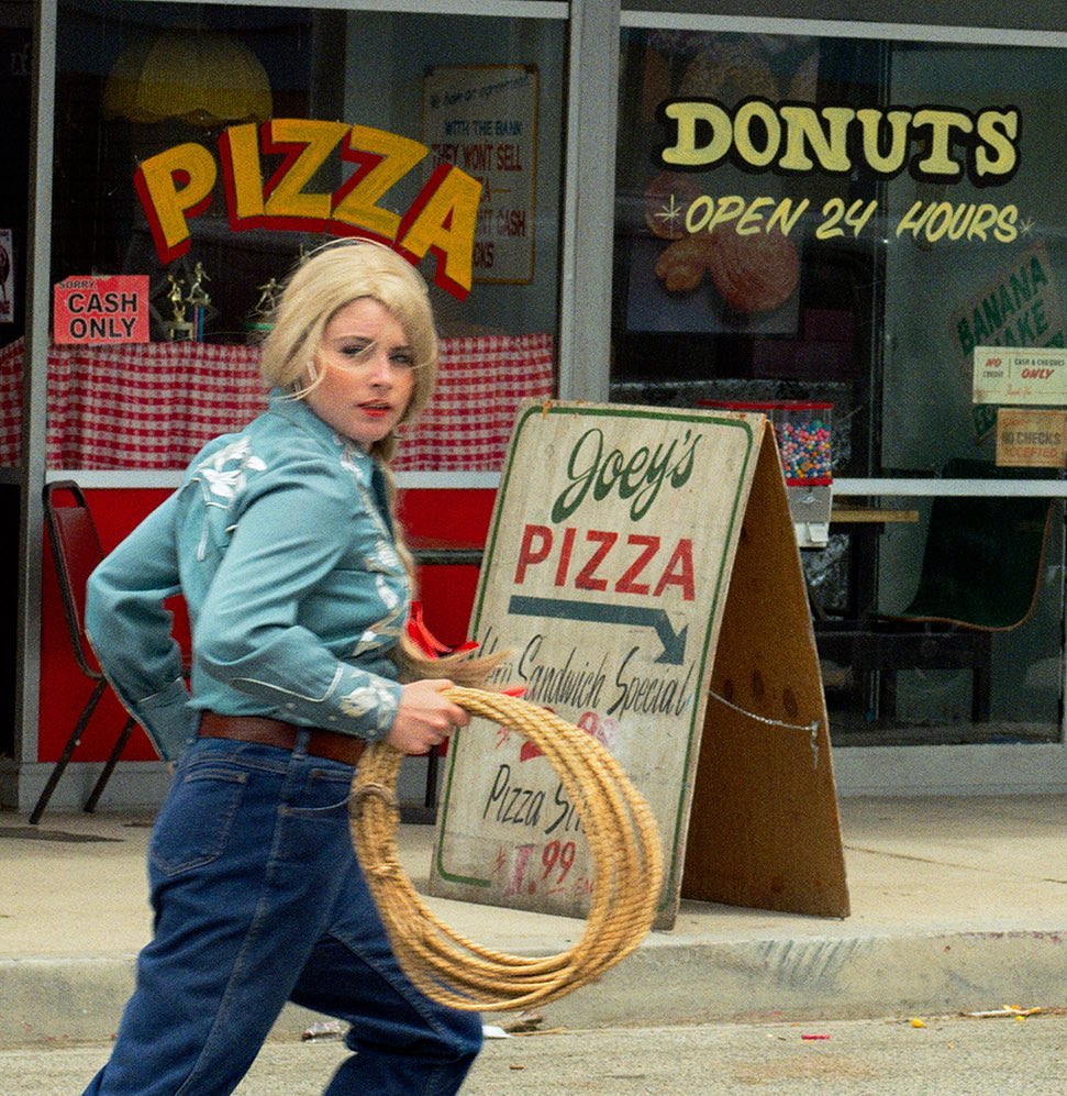 Woman in denim with a rope, walking past Joey's Pizza and a donut shop with visible signage, set outdoors in daylight.