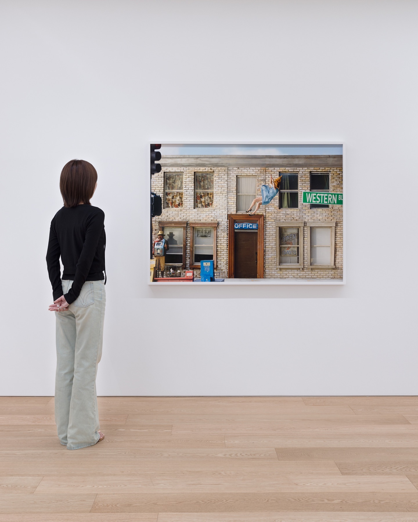 Person viewing photograph of building exterior with woman on roof and signage that reads 'Western' in a museum setting with wood floors.
