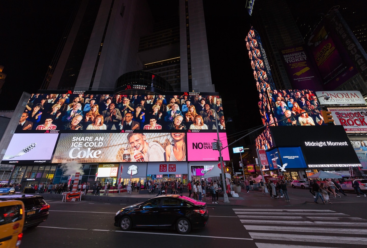Times Square at night featuring large billboards displaying ads for Coke and various other companies, with many people on the sidewalks.