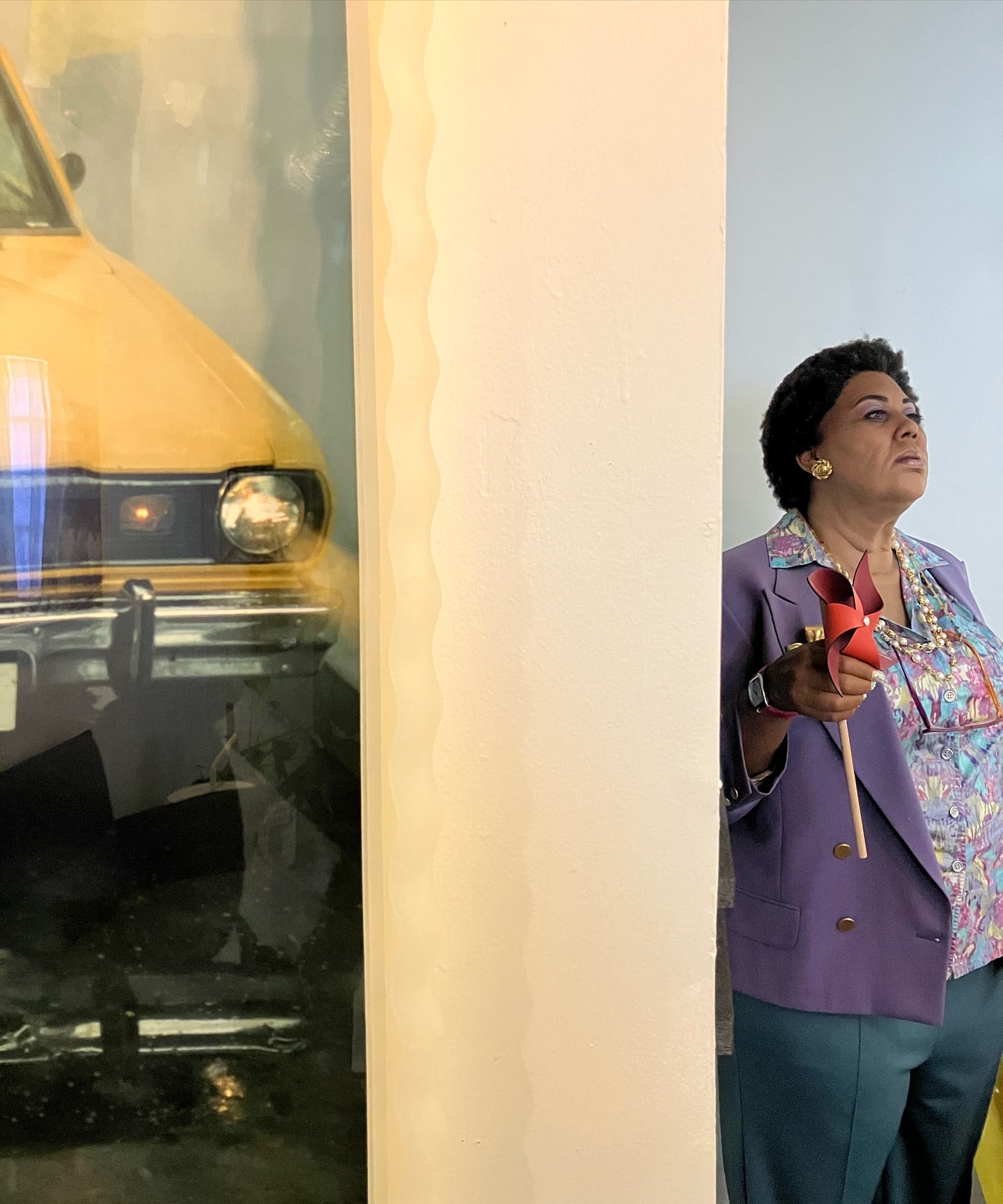 An older adult female is holding a red toy pinwheel, standing next to a wall and a painting of a yellow car in an indoor setting.