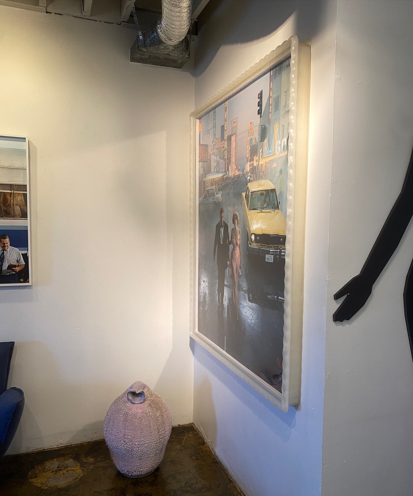 Artwork depicting an urban scene with a yellow car and two figures walking, complemented by a decorative vase in an interior setting.