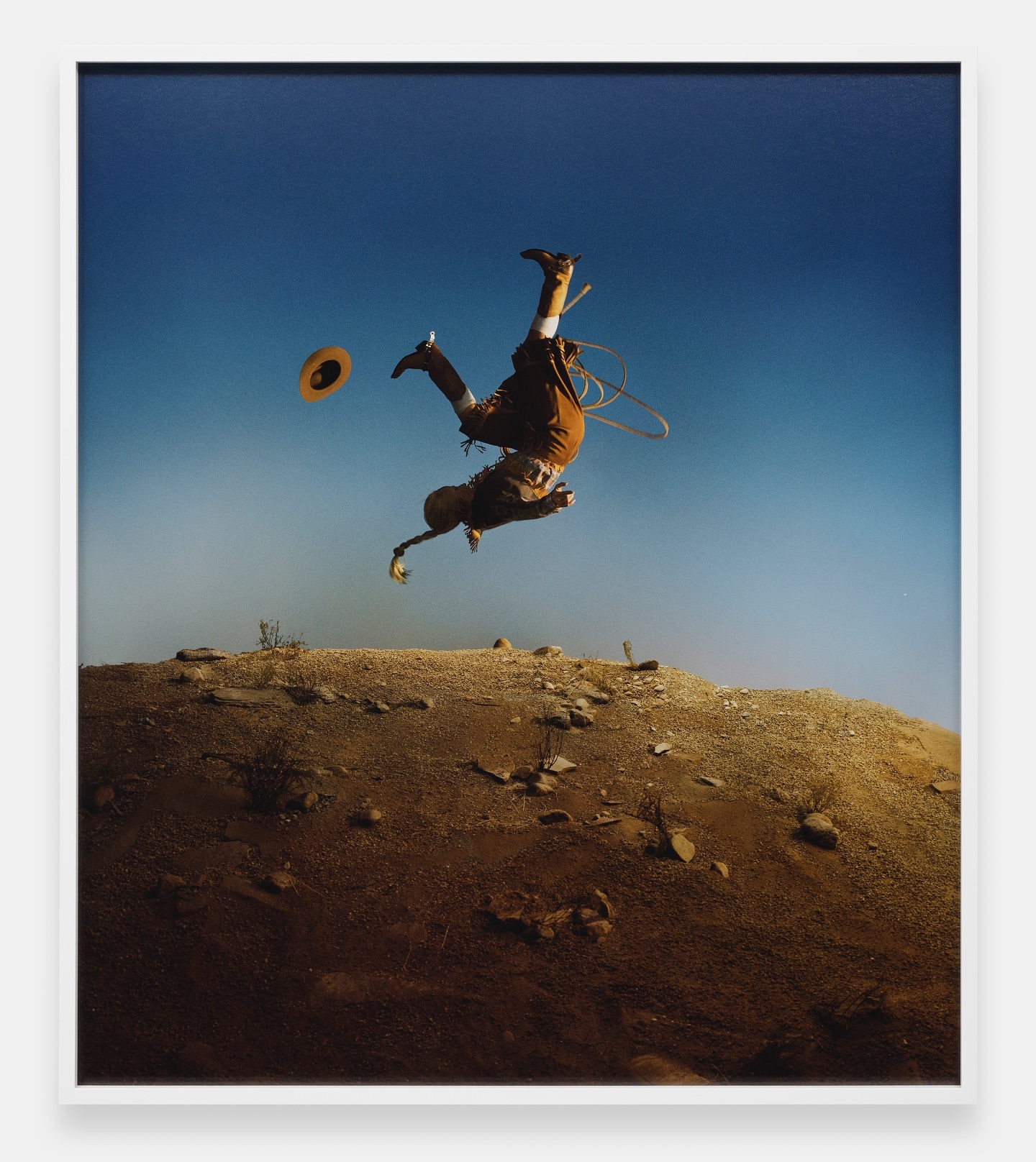 A cowboy in mid-air performing a stunt, hat floating above, set against a clear blue sky and desert landscape.