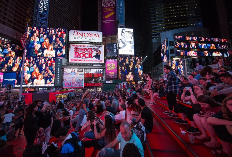 Nighttime crowd of people sitting on red steps at Times Square, New York City, surrounded by illuminated advertising billboards.