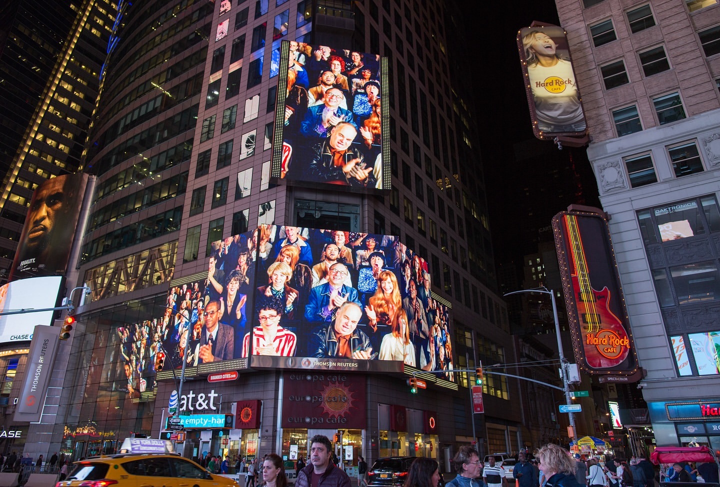 A vibrant Times Square at night featuring a large LED screen displaying a crowd of people, with city buildings and street activity.