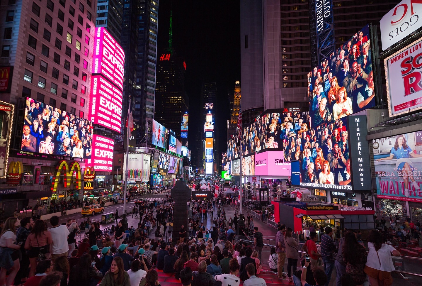 Nighttime view of Times Square in New York City with crowds, illuminated billboards, and bright lights on buildings, creating an urban scene.