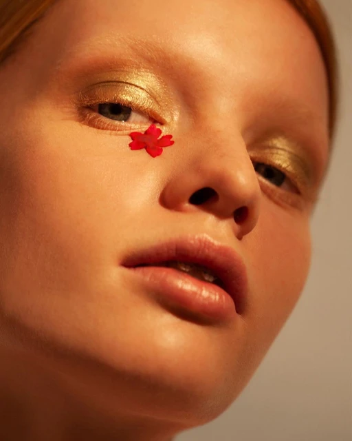 Close-up portrait of a fair-skinned adult female with a small red flower by her eye, gold eye makeup, and an orange skin tone.